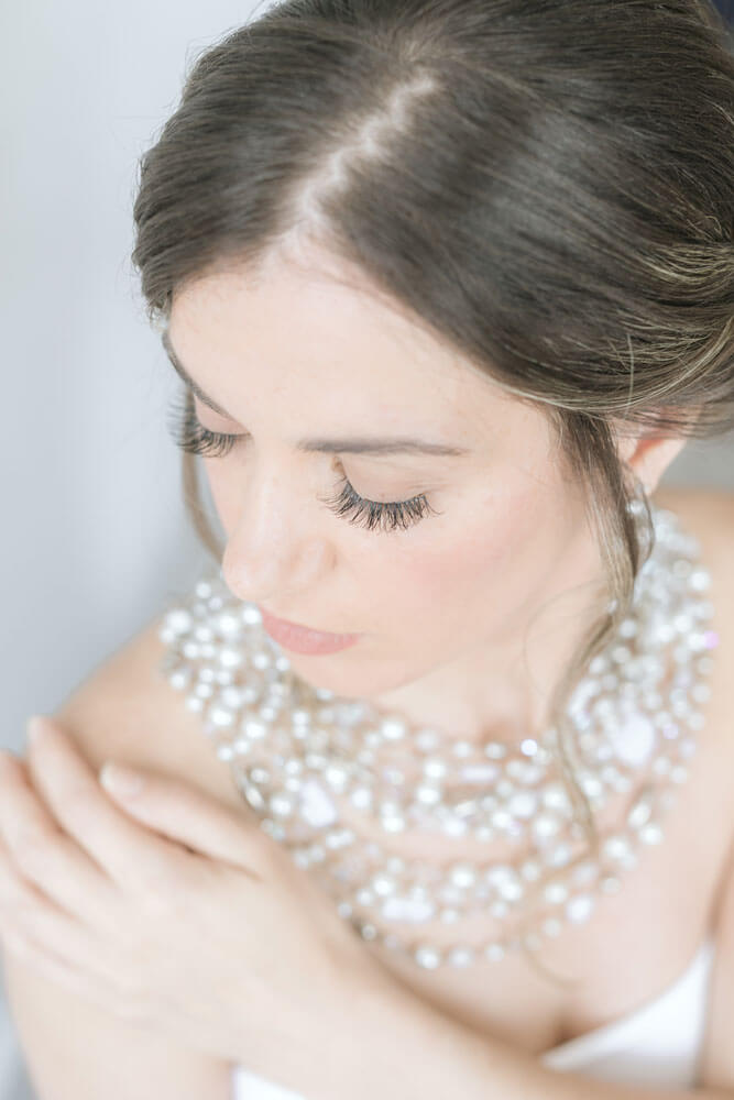 Bride wearing large shimmering neckpiece and natural fluffy lashes looking down over her shoulder.