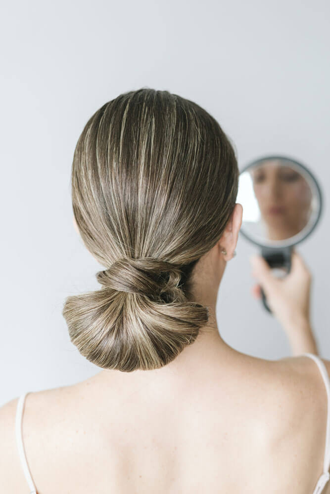Ash-blonde bride with sleek low knot bun looking into a handheld mirror.
