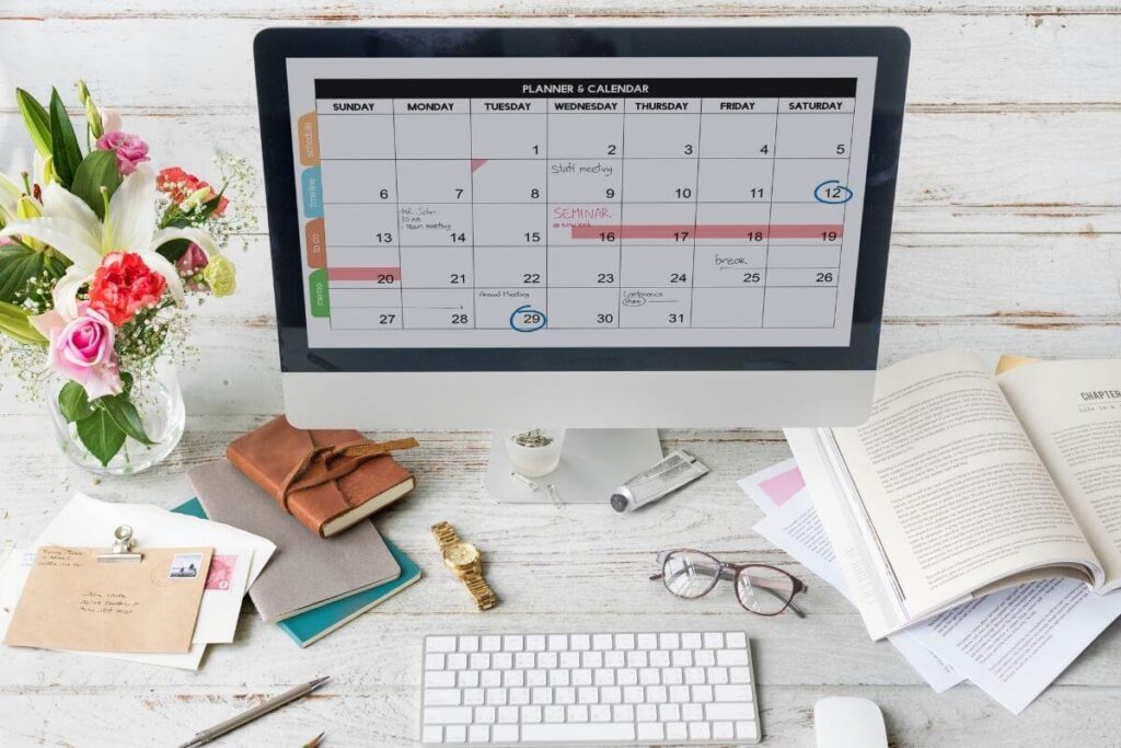 Planner and calendar on a mac computer. White wood desk with glasses, flowers, book, letters, watch, hand lotion and stationary.