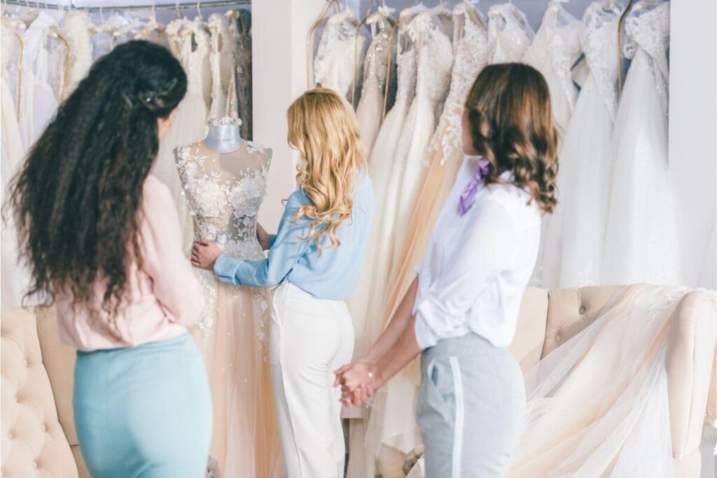 Bride with long blonde curled hair standing with long dark-haired and short copper-haired bridesmaids surrounded by bridal gowns.