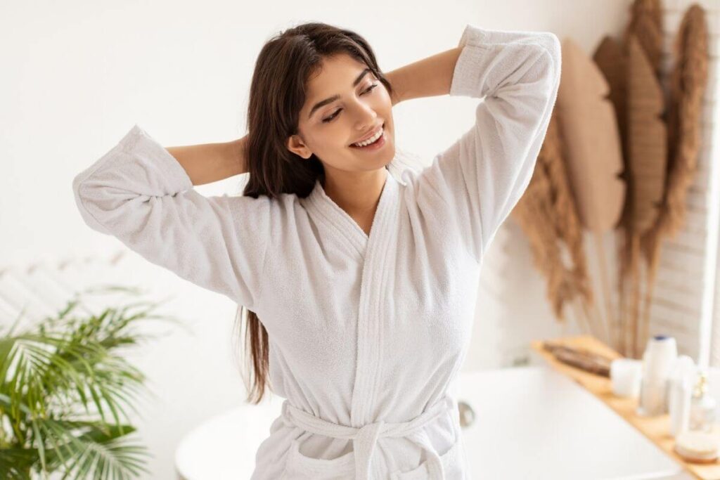 Woman with soft natural makeup and long brown hair wearing a white robe smiling and stretching in a white aesthetic bathroom.