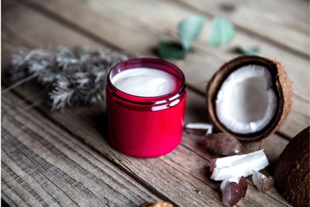 Red glass tub filled with cream on a wooden table next to a half coconut and sage leaves