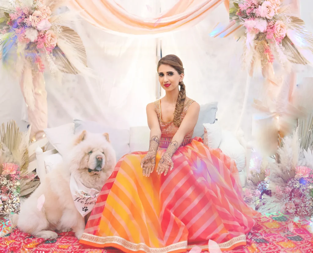 Bride in vibrant orange and pink lehenga with henna designs, sitting with her dog under pastel floral décor