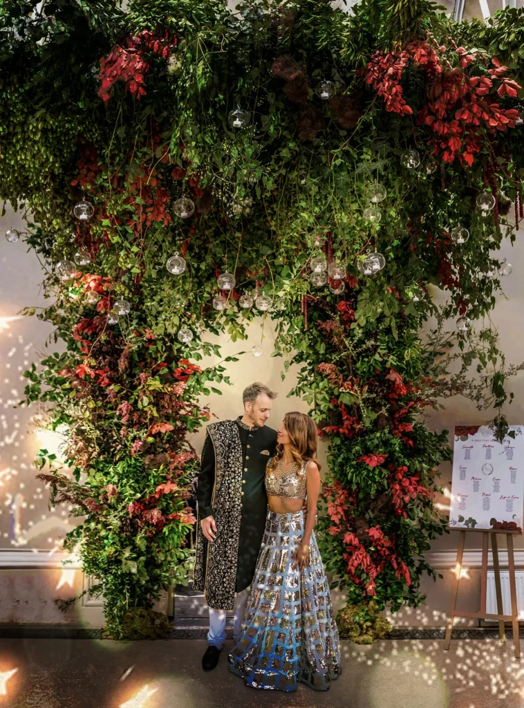 Couple in traditional South Asian attire standing under a dramatic floral arch with red and green foliage