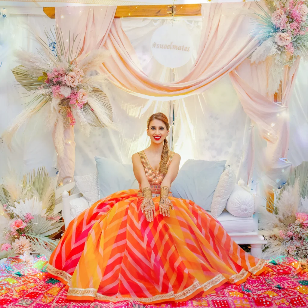 Bride in colorful lehenga with henna on her hands, seated under pink draped canopy with pastel flowers