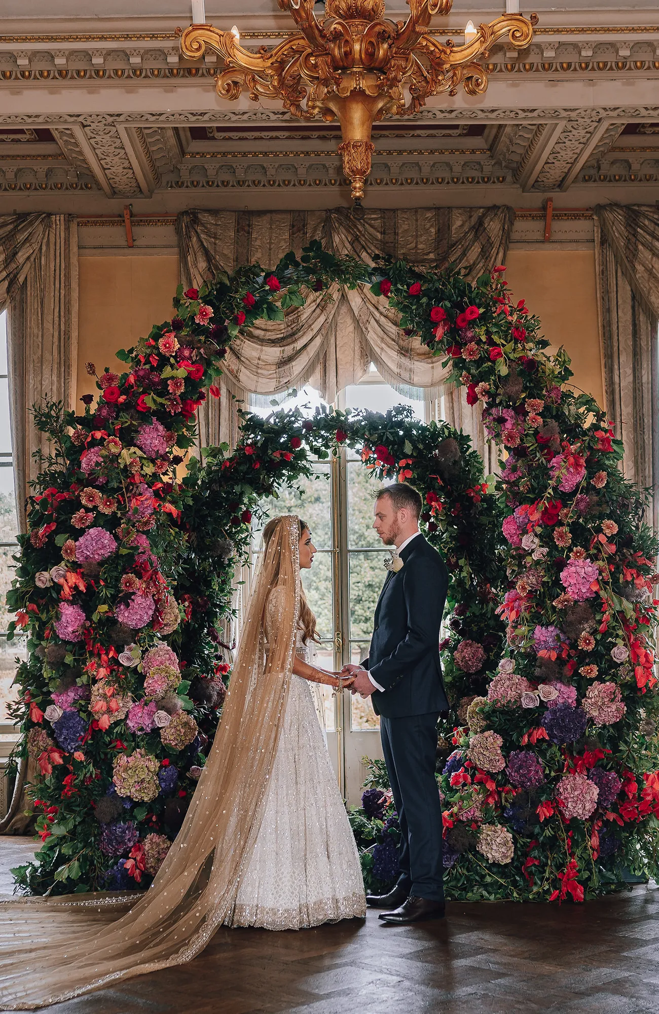 Bride and groom holding hands under a luxurious circular floral arch of roses and hydrangeas at Hampton Court House Wedding ceremony.