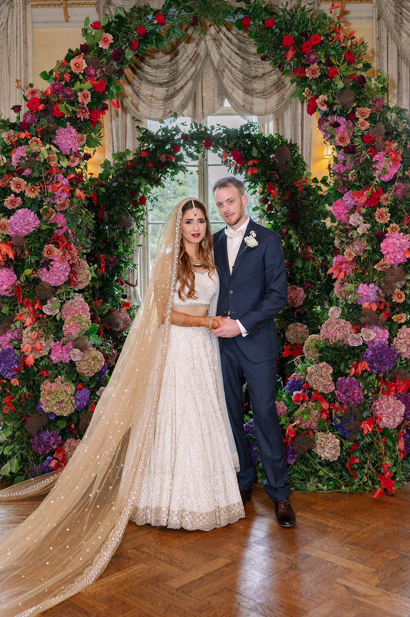 Bride and groom posing together under an elaborate floral arch inside Hampton Court House Wedding venue.