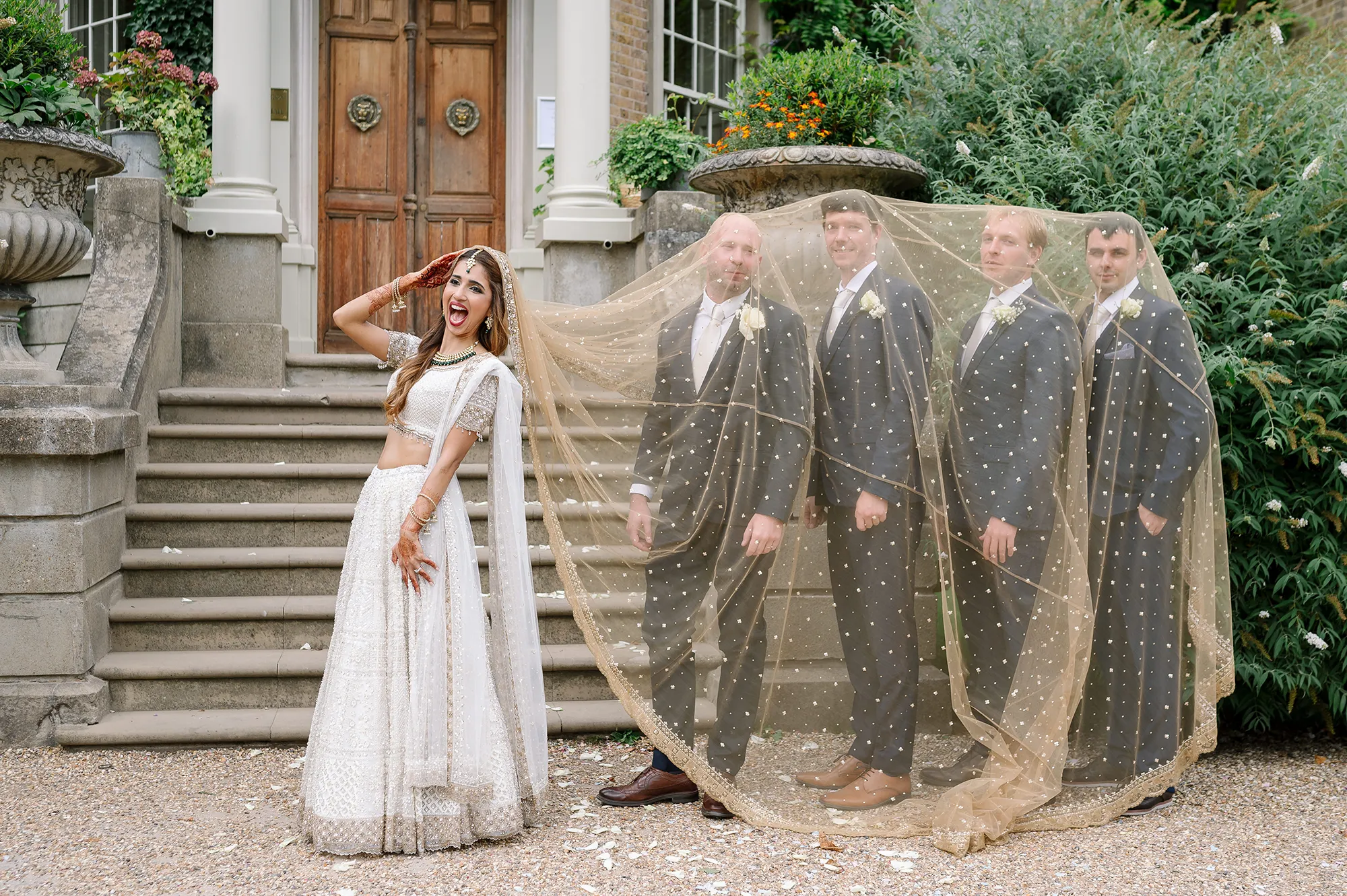 Bride playfully posing with groomsmen under her golden veil outside Hampton Court House