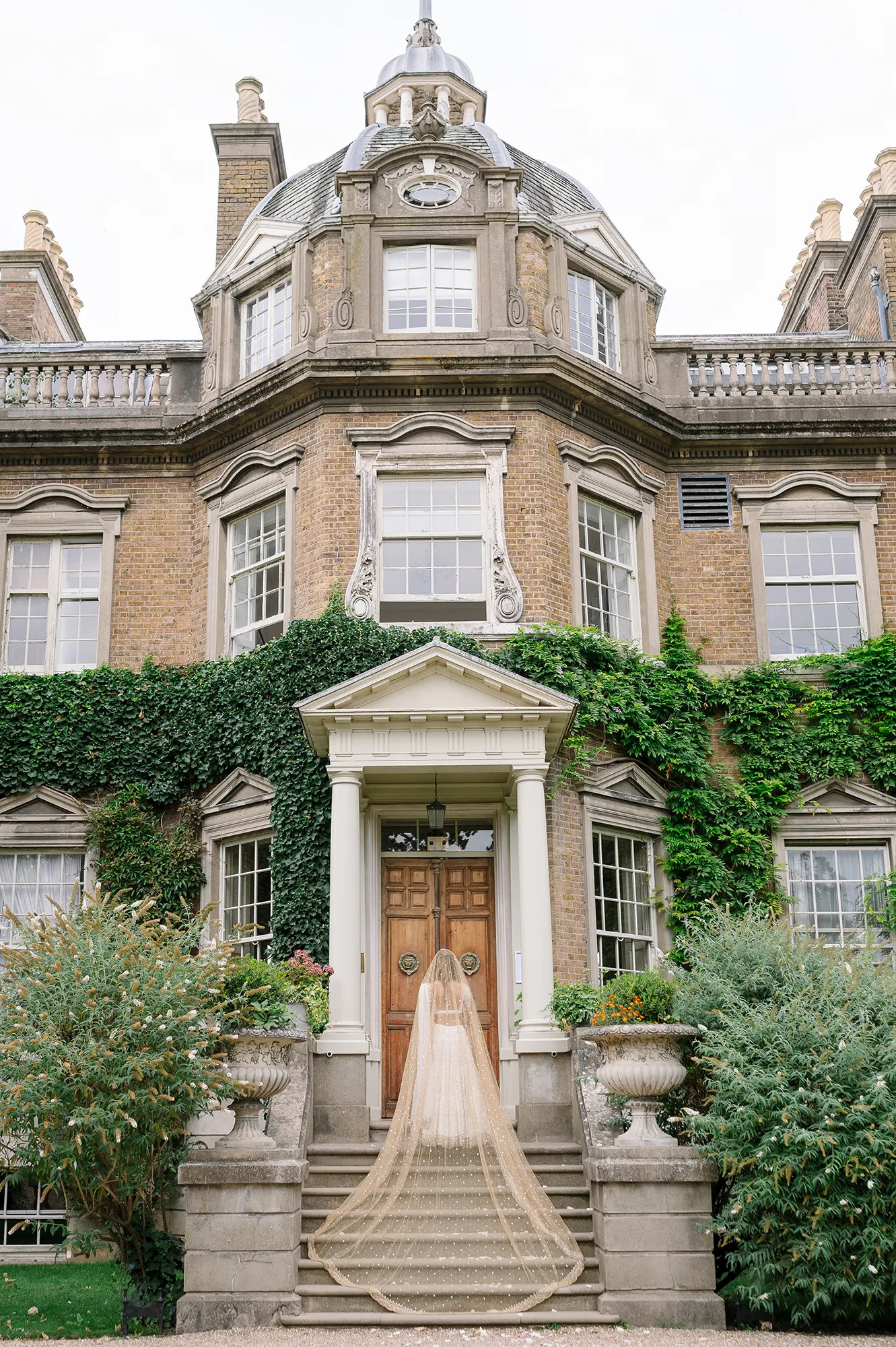 Back view of bride in flowing golden veil on the grand staircase of Hampton Court House Wedding venue