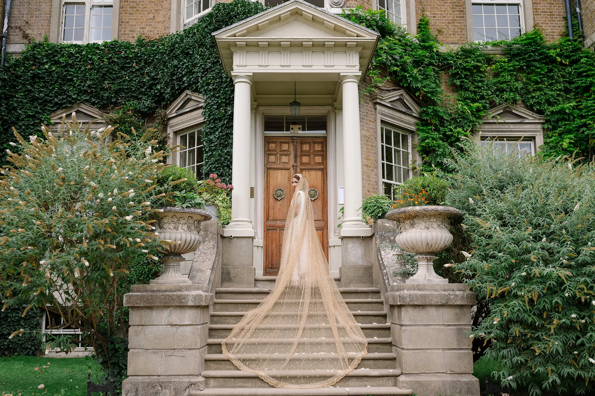 Bride standing on the grand staircase of Hampton Court House in a golden veil, showcasing the venue’s historic architecture.