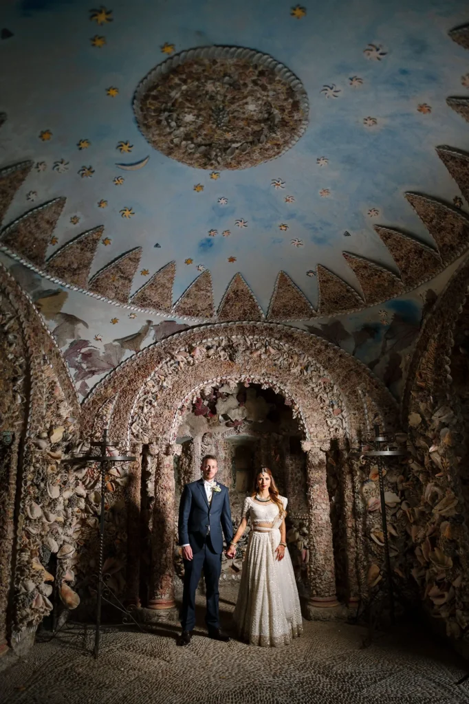 Bride and groom holding hands inside the Shell Grotto at Hampton Court House Wedding, surrounded by ornate stone and seashell walls.