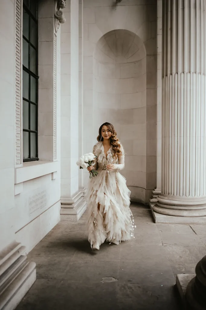 Bride Dannah in a ruffled dress holding flowers at a columned Historic London Old Marylebone Town Hall.