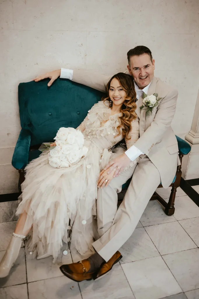 Bride Dannah and groom David seated on a teal velvet sofa at The Old Marylebone Town Hall in London.
