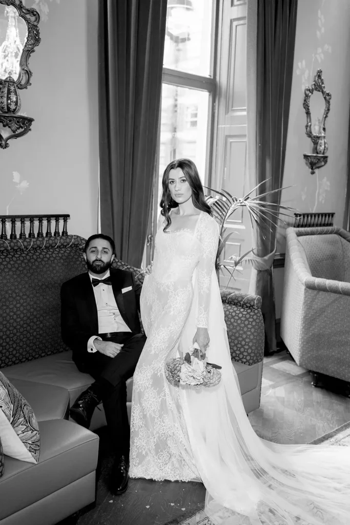 A black and white photo of a bride and groom in an elegant room. The groom is sitting on a sofa wearing a black tuxedo, while the bride stands beside him in a long, lace wedding gown with a long train. The room features ornate mirrors and draped curtains.