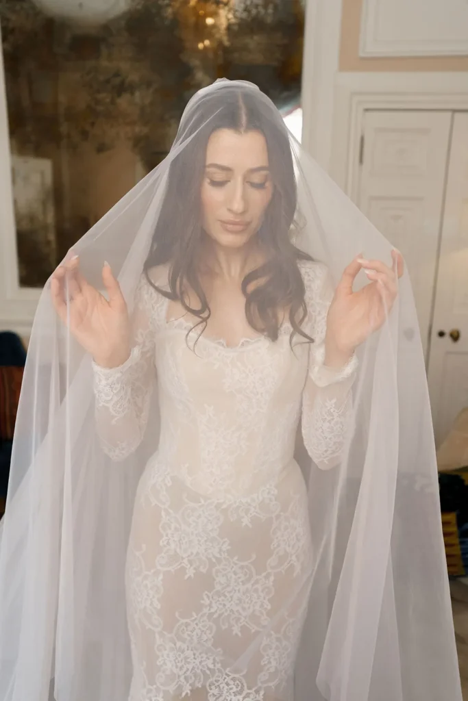 A close-up colour photo of a bride with long, dark hair, covered by a sheer veil. She wears a lace wedding dress and is looking down, holding the veil with both hands.