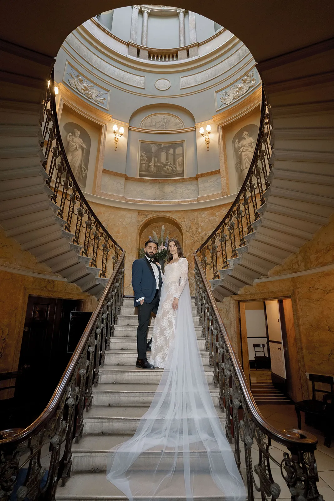 The bride and groom are standing on a grand staircase inside Home House London. The bride wears a long lace dress with a flowing train, and the groom is in a dark suit. The staircase has intricate railings, and the backdrop includes classical architectural details.