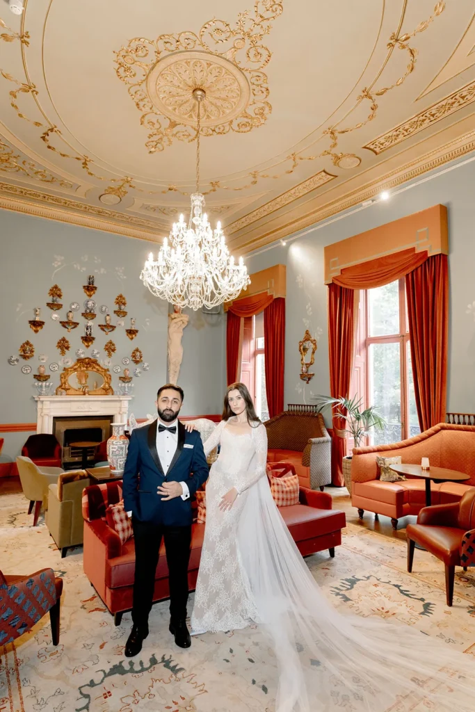 The couple is posing in a luxurious room with high ceilings and chandeliers. The groom is wearing a blue tuxedo, and the bride is in a lace wedding dress with a long train. The room has plush seating, ornate wall decorations, and large windows with heavy drapes.