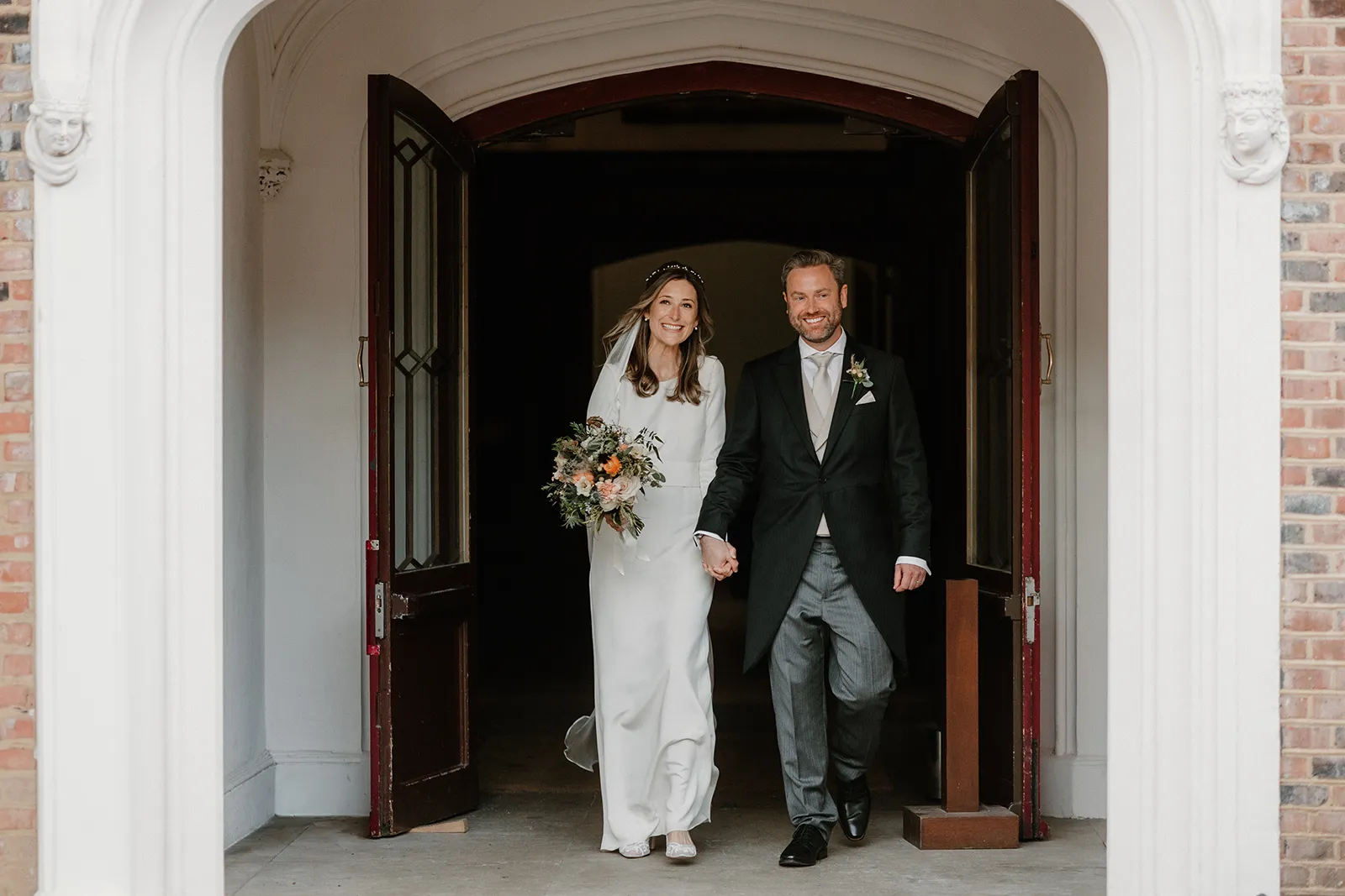 Bride and Groom, the newlyweds, holding hands and smiling as they walk out of the grand doorway at Fulham Palace, London.