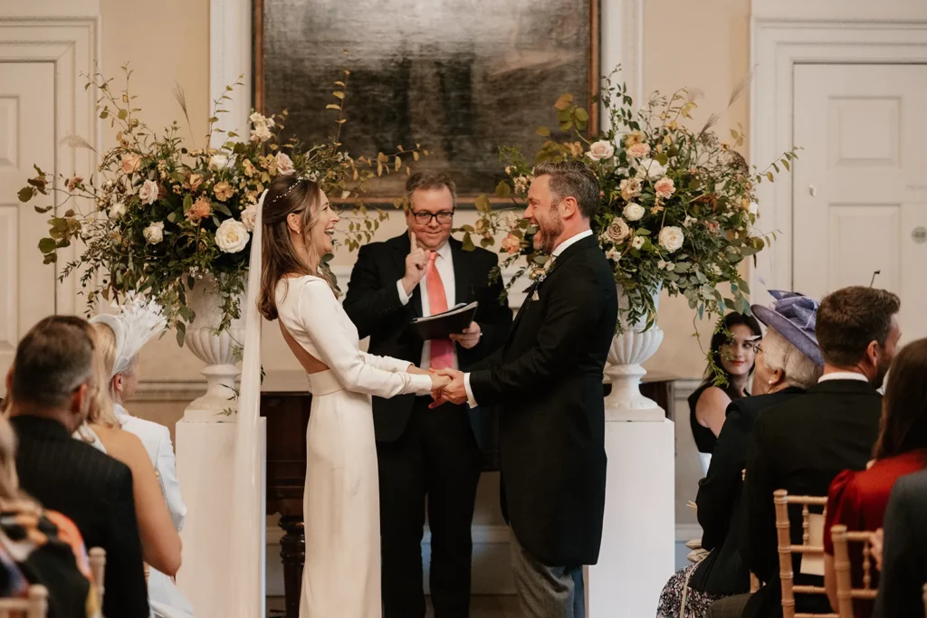 Bride and Groom standing at the altar during their wedding ceremony at Fulham Palace London, both laughing and holding hands amidst a beautifully decorated setting.