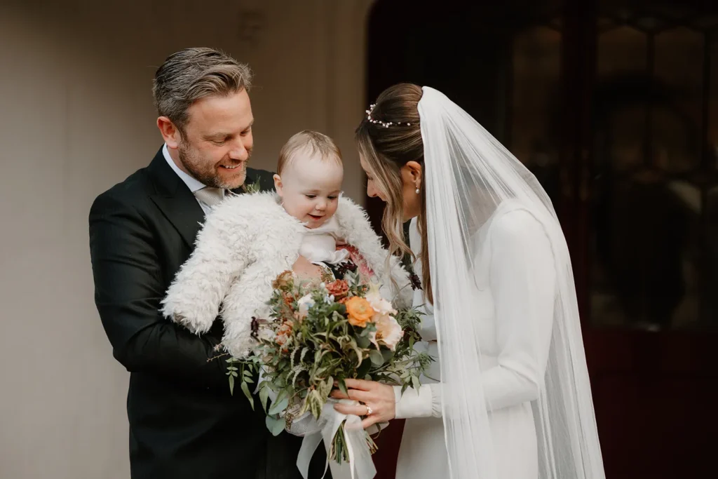 Bride and Groom smiling warmly as they hold their baby outside Fulham Palace London, with Isabella gently showing the baby her wedding bouquet.