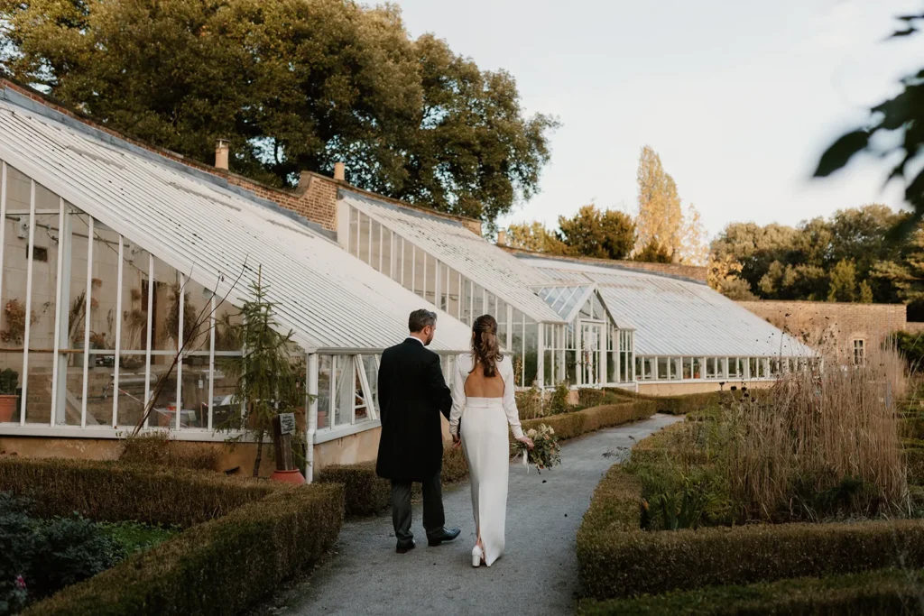 Bride and Groom walking hand in hand through the botanical gardens at Fulham Palace, with the beautiful glass greenhouses in the background.