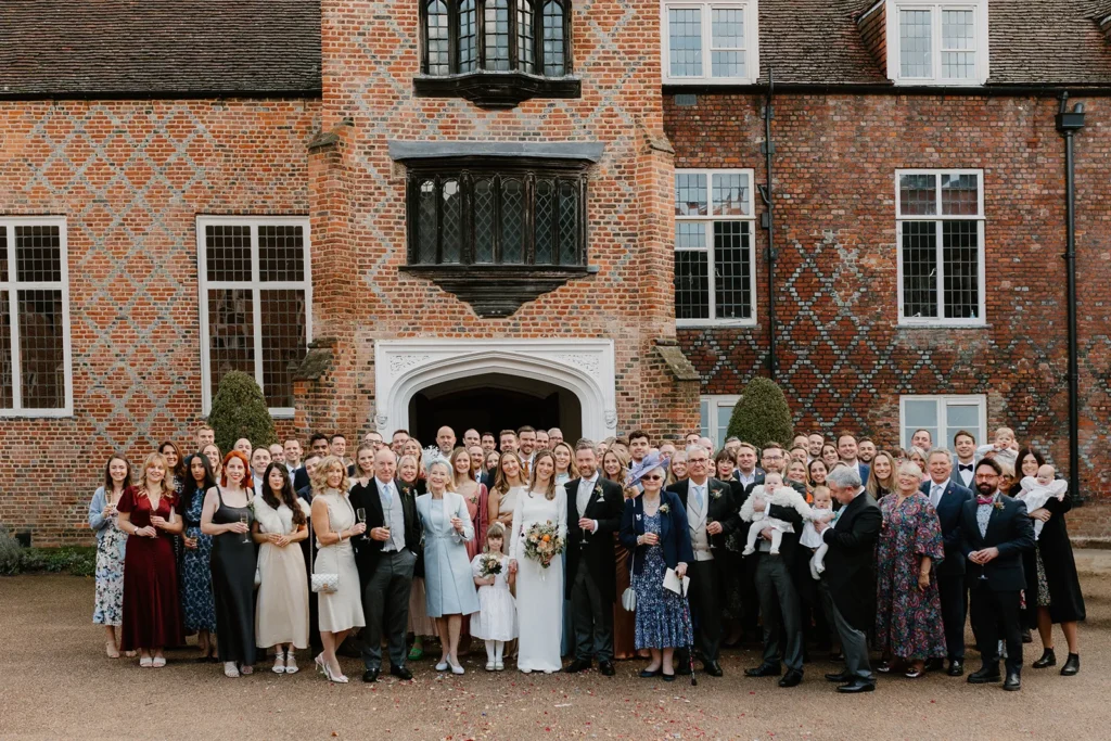 A large group of wedding guests posing in front of the historic brick facade of Fulham Palace London, smiling and celebrating Isabella and Adrian's special day.