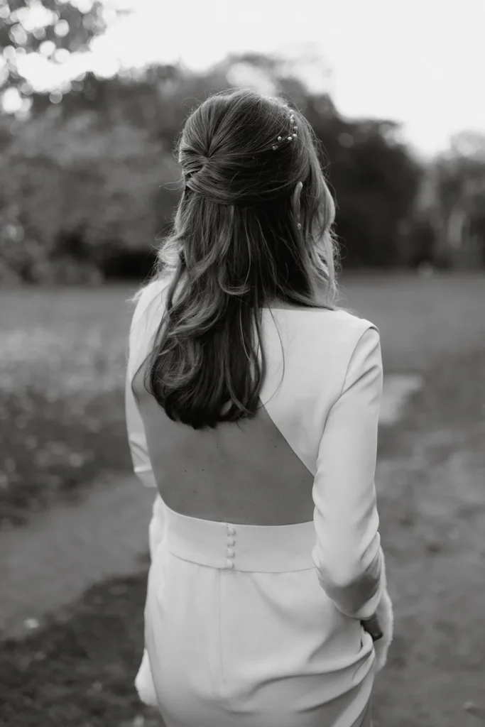 A black and white image capturing the back of Isabella in her wedding dress at Fulham Palace, highlighting her elegant hairstyle adorned with delicate pearls and the stylish open-back design of her gown as she stands in the garden.