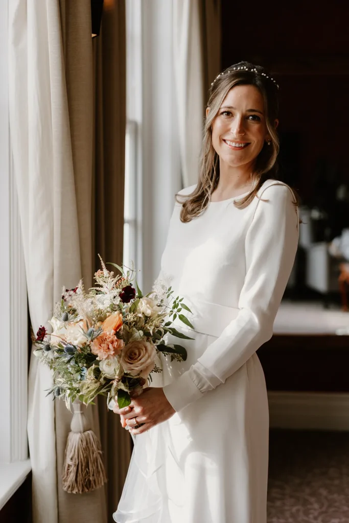 A radiant bride, standing by a window at Fulham Palace London, holding her exquisite wedding bouquet filled with soft pink and peach flowers.