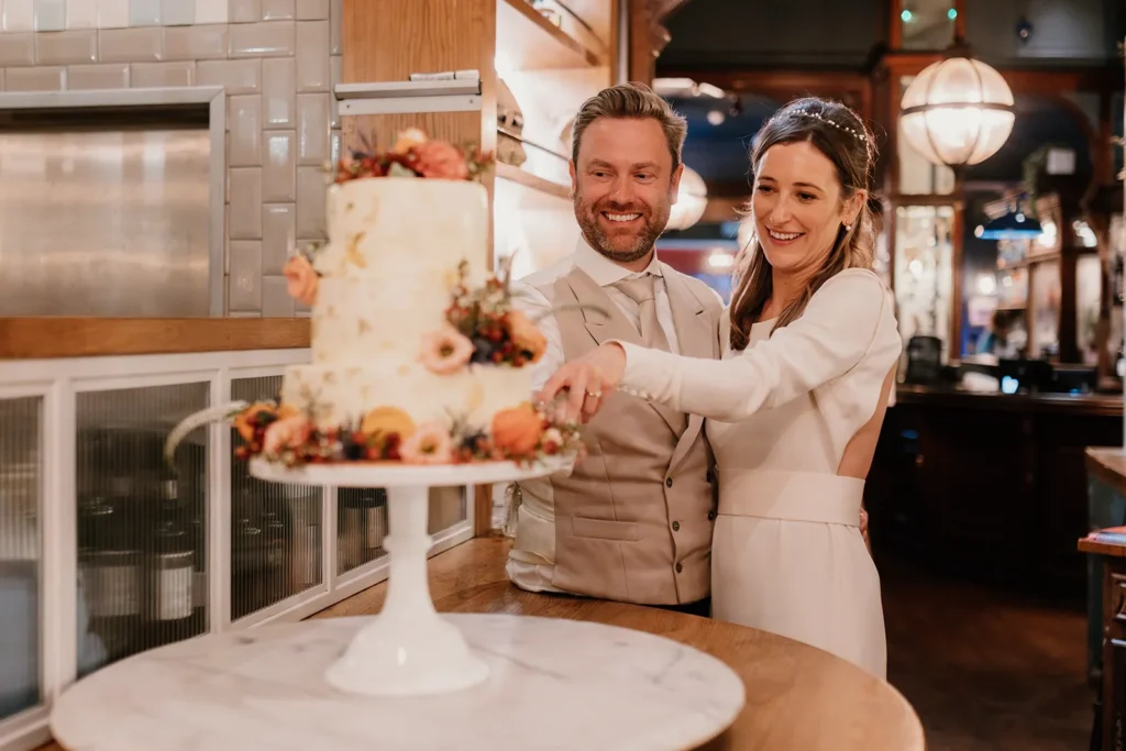 Bride and Groom smiling as they cut their elegantly decorated wedding cake at Fulham Palace London, surrounded by warm lighting and joyful ambiance.