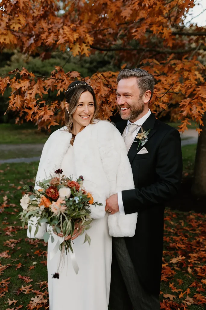 Bride and Groom holding a vibrant wedding bouquet, standing under a tree with autumn leaves at Fulham Palace London, sharing a joyful moment.