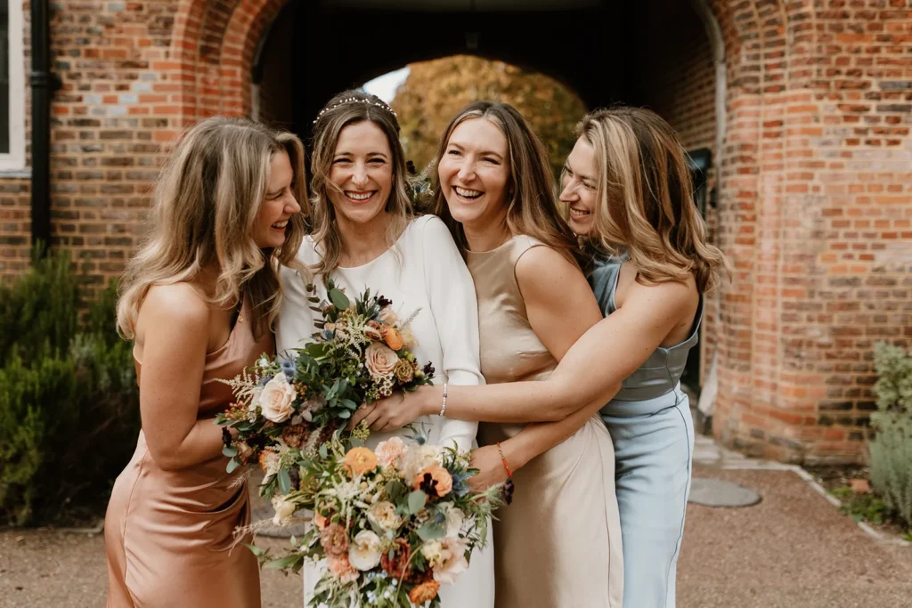 A candid moment of Isabella, the bride, laughing and hugging her bridesmaids, all holding bouquets and radiating happiness outside Fulham Palace London.