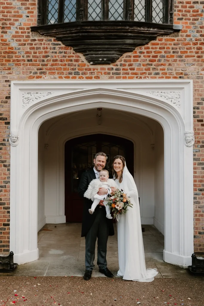 Bride and Groom posing with their adorable baby outside Fulham Palace London, the bride holding her bouquet, all smiling warmly at the camera.