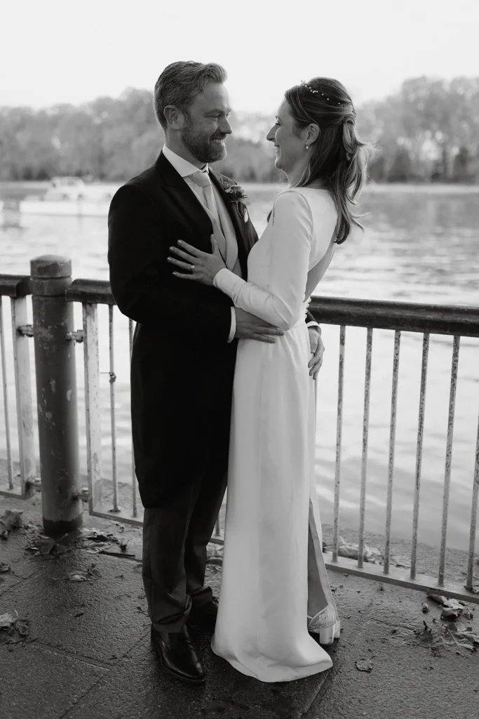 A black and white photo of Isabella and Adrian standing close by the river at Fulham Palace London, smiling at each other with deep affection.