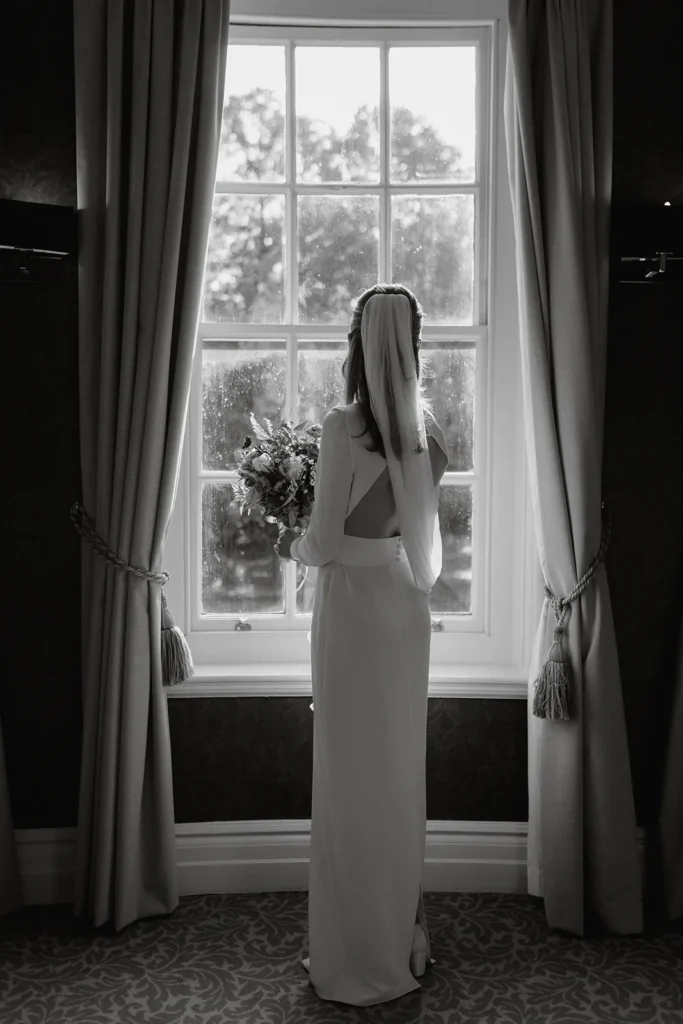 The bride, standing alone and looking out through a large window at Fulham Palace London, holding her bouquet, with sunlight streaming in.