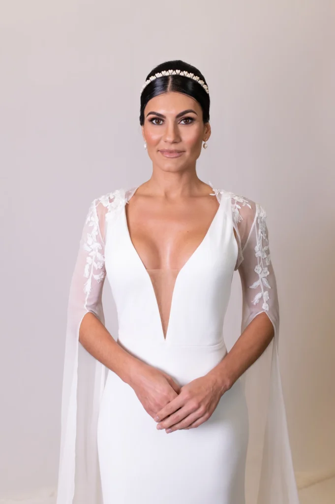 Bride in white gown with sheer lace sleeves, pearl headband, and sleek low bun hairstyle