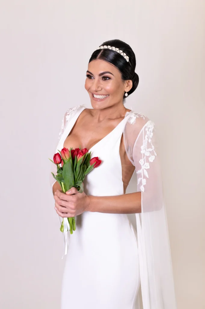 Bride posing in an elegant white dress with a low back and cape, holding red tulips