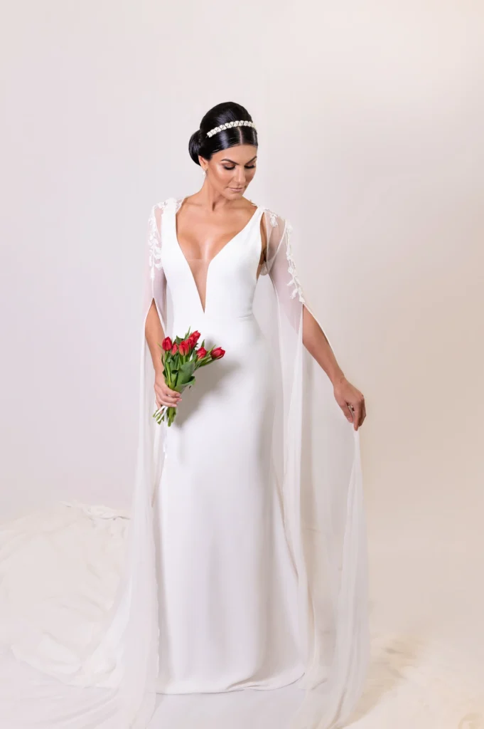 Bride standing confidently in a plunging white gown with sheer floral-embroidered cape