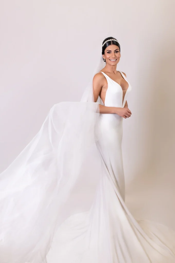 Bride in a sleek white gown with flowing veil, smiling elegantly for the camera