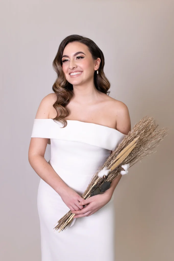 Smiling bride in off-shoulder white gown holding a bouquet of dried wheat