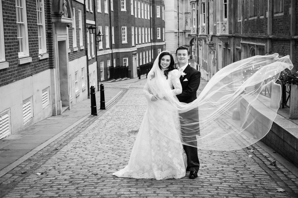 Bride and groom captured in an intimate embrace on a cobblestone street, veil flowing in the winter breeze.