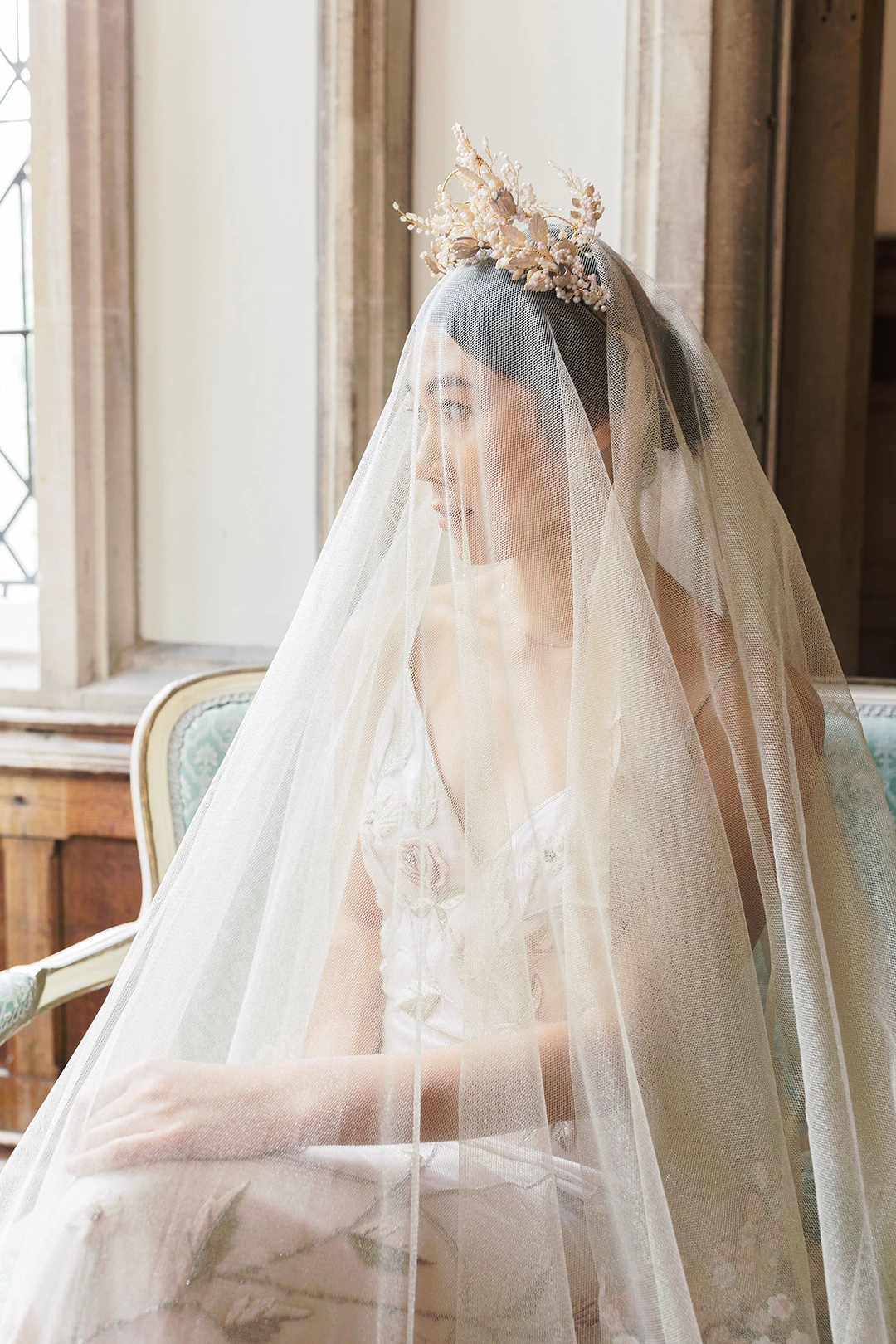 Bride in an ethereal veil and floral crown seated by a window at Nutfield Priory.