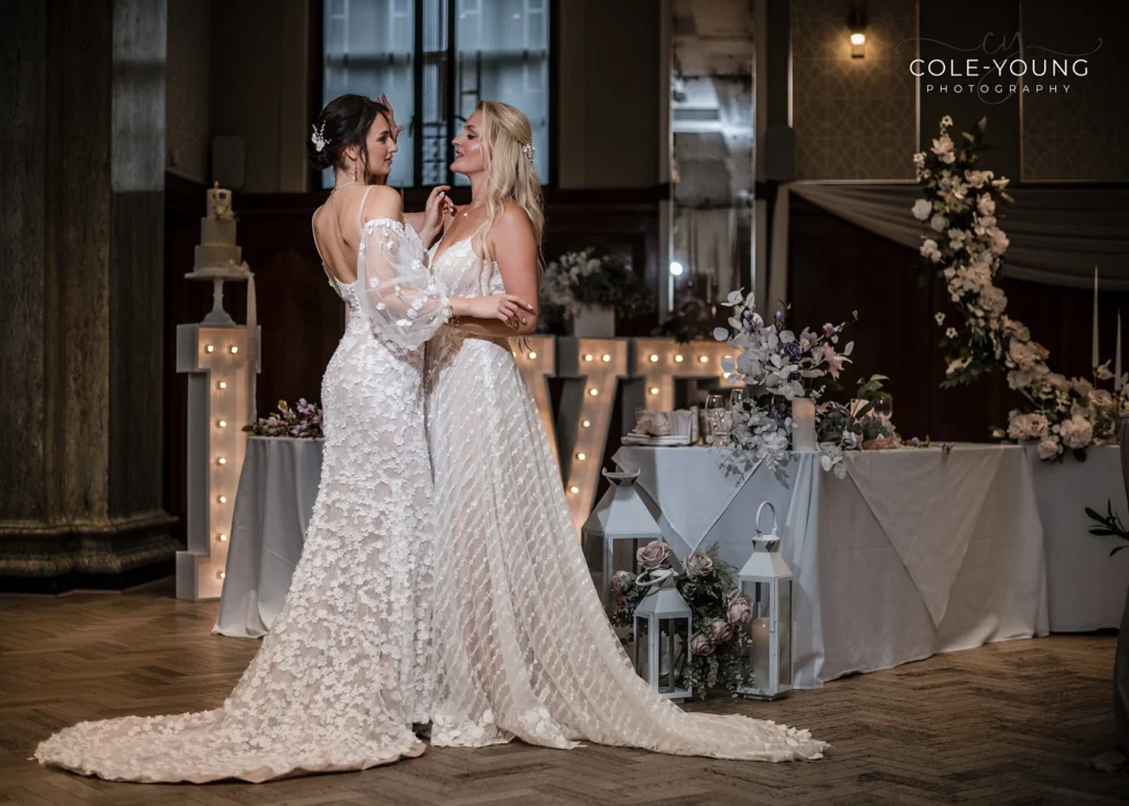 Two brides embracing in detailed wedding dresses during a Pelham House celebration.