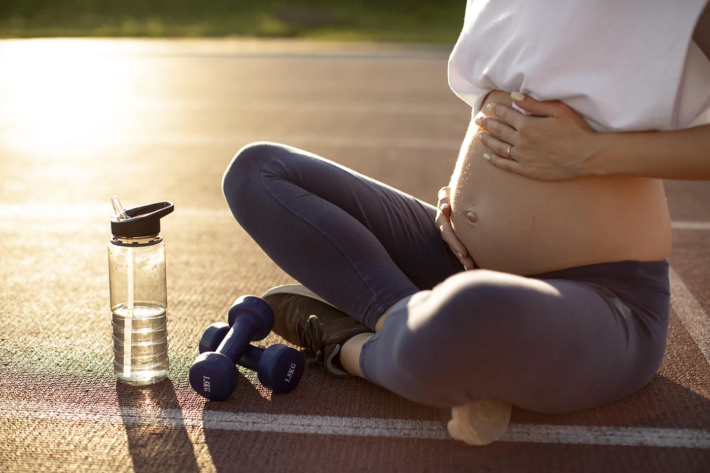 Pregnant woman sitting outdoors with water bottle and dumbbells