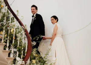 Bride and groom walking hand in hand up a grand staircase adorned with lush greenery and white florals at their elegant wedding