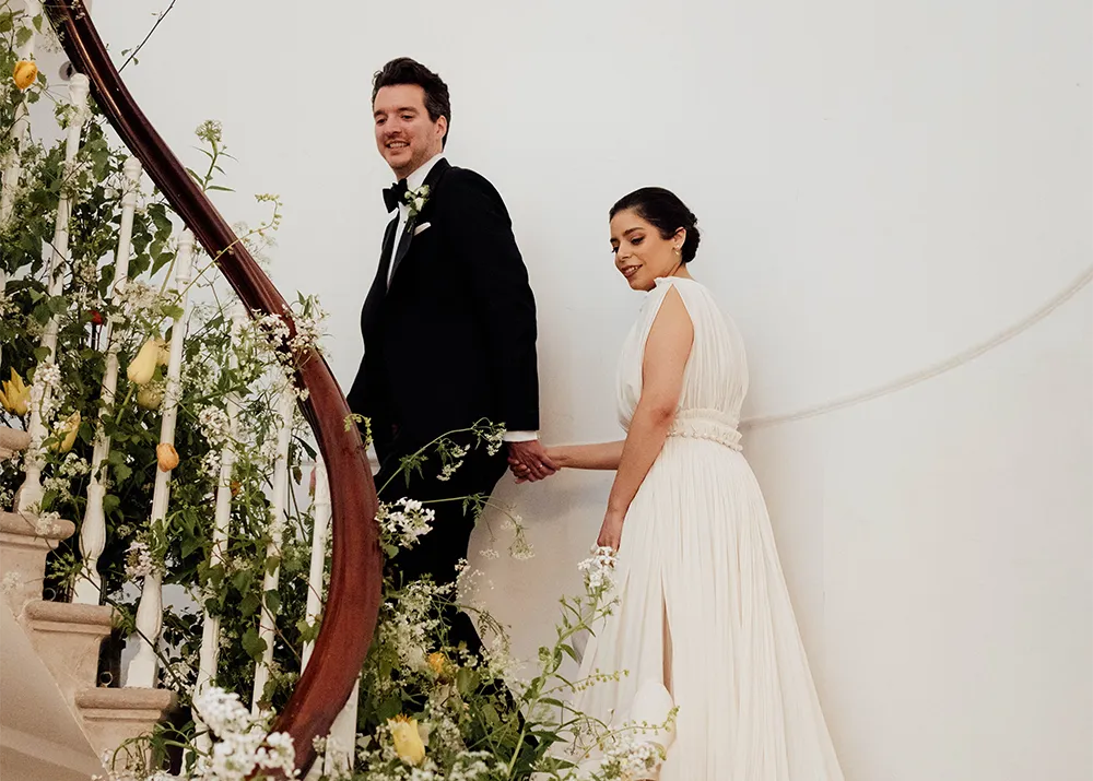 Bride and groom walking hand in hand up a grand staircase adorned with lush greenery and white florals at their elegant wedding