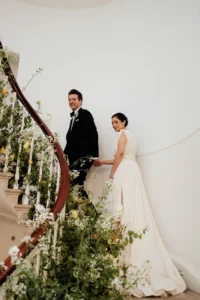 Bride and groom walking hand in hand up a grand staircase adorned with lush greenery and white florals at their elegant wedding