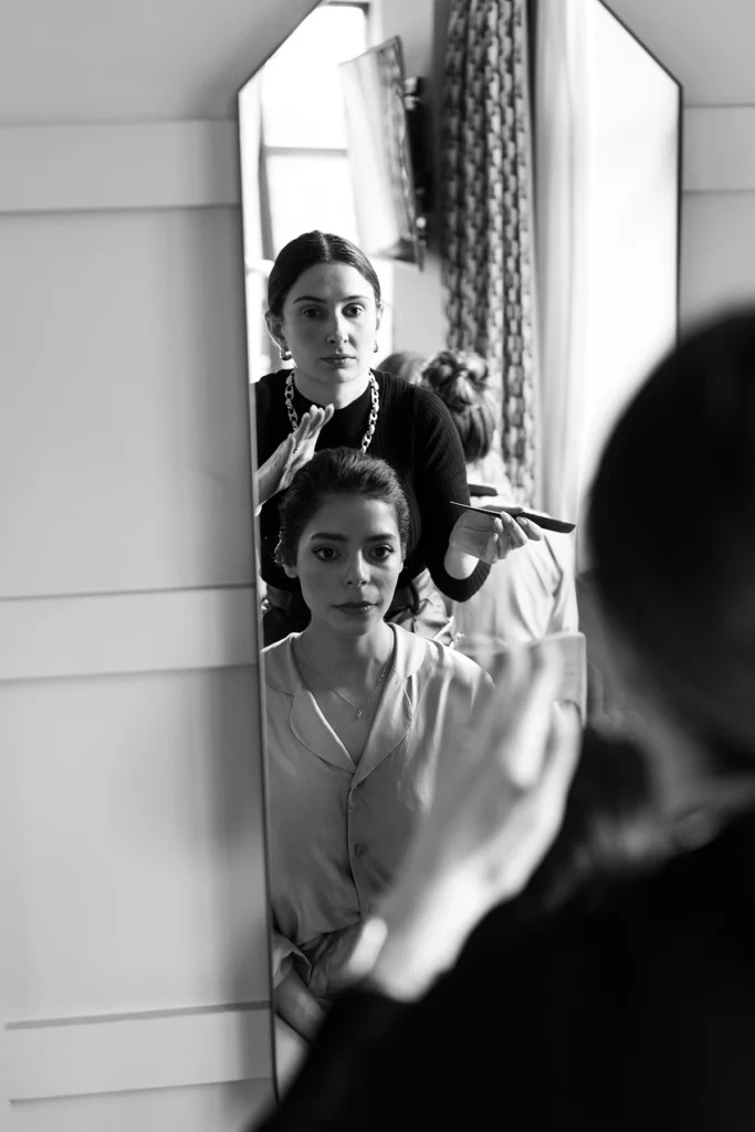 Bride looking at her reflection in the mirror while her hairstylist carefully finishes her elegant wedding updo