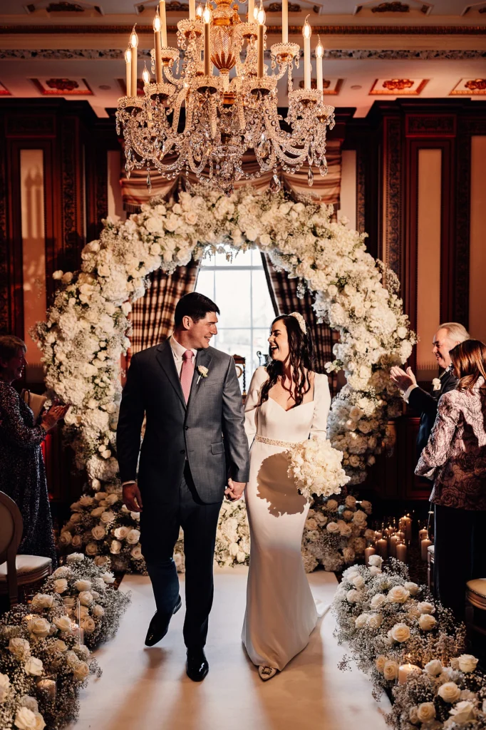 Bride and groom walking down the aisle under a chandelier and floral arch