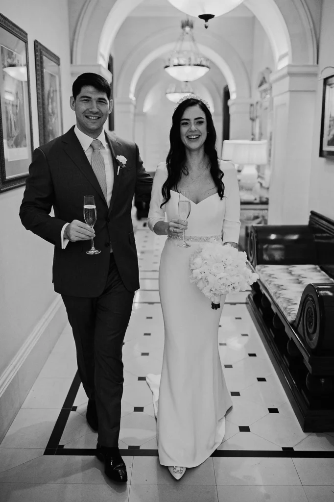 Bride and groom walking with champagne glasses in hand inside Lanesborough Hotel