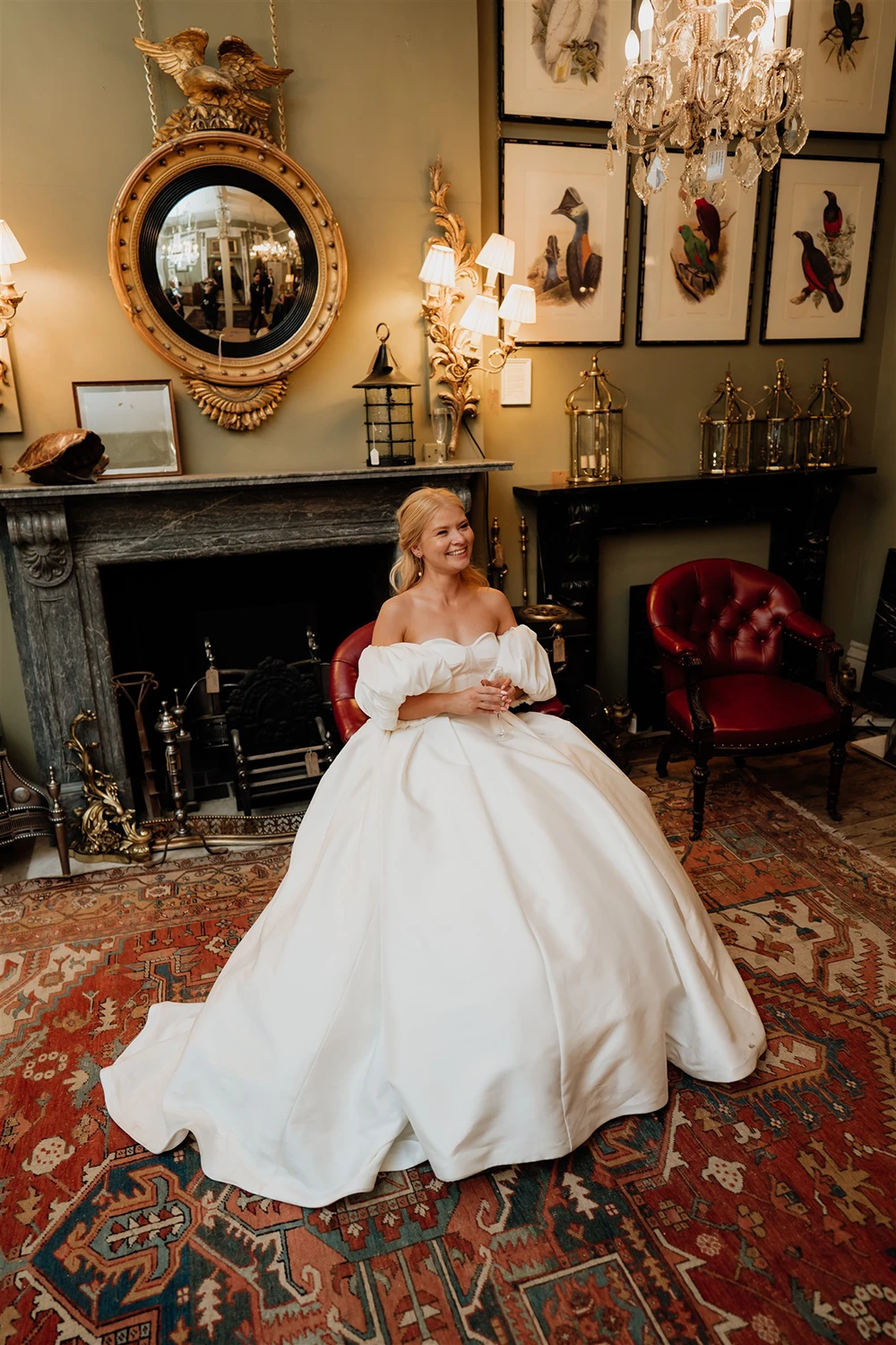 Bride sitting in an ornate room wearing a full wedding gown surrounded by framed artwork and chandeliers at Brunswick House
